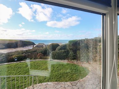 A view of a garden with grass and a stone path looking out to the sea and nearby houses at Gwillen in Mawgan Porth