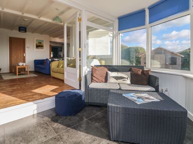 A sunroom with wicker sofa and ottoman with cushions next to a living room with blue and yellow sofas at Gwillen in Mawgan Porth