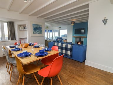 A dining area with a wooden table set with blue plates and colorful chairs next to a living room with a blue sofa and a fireplace at Gwillen in Mawgan Porth