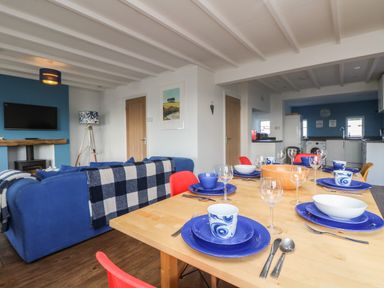 A dining table set with blue plates and cups next to a blue sofa and a kitchen in the background at Gwillen in Mawgan Porth
