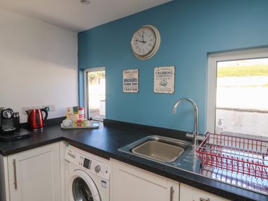 A kitchen corner with a washing machine under counter a sink with faucet and a red dish rack tea and coffee items on black countertop and a blue wall with a clock and two nautical themed signs at Gwillen in Mawgan Porth