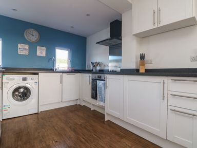 A kitchen with white cabinetry wooden floor washing machine stove and blue wall at Gwillen in Mawgan Porth