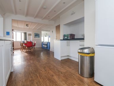 A kitchen area with a white refrigerator a stainless steel trash can and white cabinets leading to a dining area with a round table red and beige chairs at Gwillen in Mawgan Porth