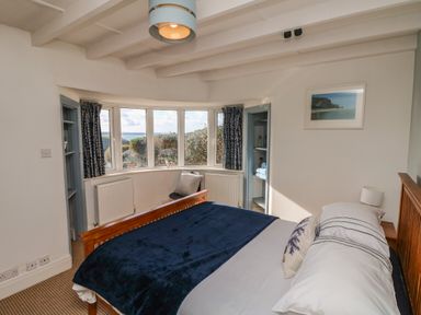 A bedroom with a wooden bed and blue blanket near a bay window with curtains at Gwillen in Mawgan Porth