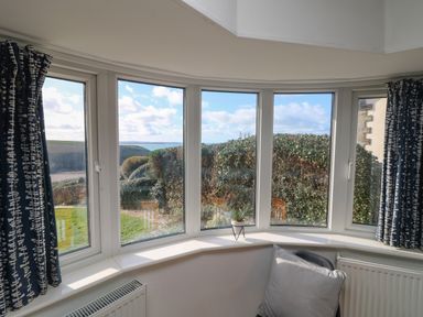 A bay window with blue patterned curtains a small plant a chair with cushions and a view of bushes and the sea at Gwillen in Mawgan Porth