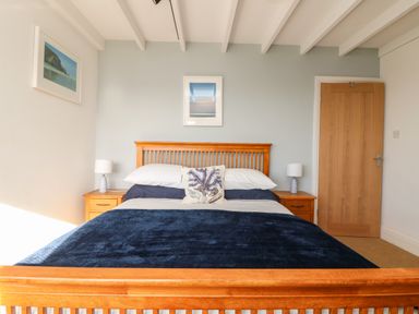 A bedroom with a wooden bed frame blue bedding two nightstands with lamps and framed pictures on walls at Gwillen in Mawgan Porth