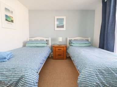 A bedroom with two single beds a wooden nightstand with a lamp and framed pictures on the walls at Gwillen in Mawgan Porth