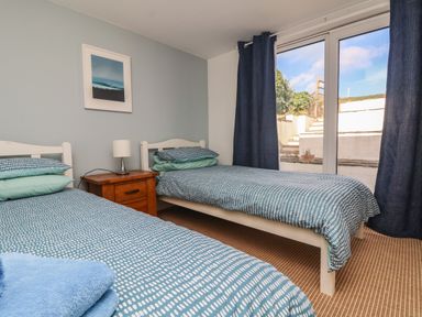 A bedroom with two single beds a wooden nightstand with a lamp and glass doors with blue curtains at Gwillen in Mawgan Porth