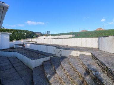 A garden with stone steps and tiered white retaining walls with grass at Gwillen in Mawgan Porth