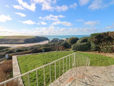 A garden with grass and stone steps overlooking houses and a beach with cliffs in the distance at Gwillen in Mawgan Porth