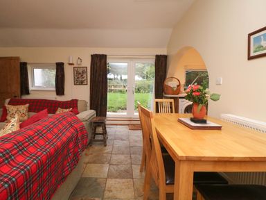A dining area with a wooden table and chairs next to a living room with sofas covered in red plaid blankets at Goslings in Bradford Holsworthy
