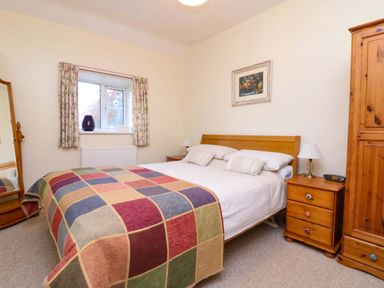 A bedroom with a double bed covered with a colorful checkered blanket a wooden wardrobe and a bedside table with a lamp and alarm clock at Goslings Bradford Holsworthy