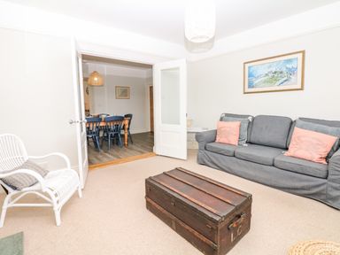 A living room with a gray sofa and coral pillows a white chair and a wooden trunk with an open door to a dining room with a wooden table and blue chairs at Cool Stone Cottage in Instow near Bideford