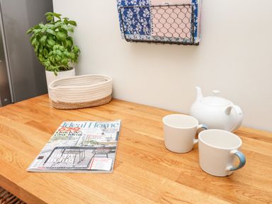 A wooden countertop with a potted basil plant a woven basket a magazine a white teapot and two white mugs with blue handles at Cool Stone Cottage Instow near Bideford