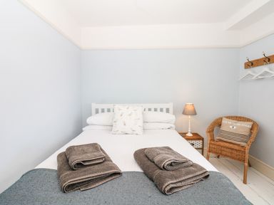 A bedroom with a white bed holding folded towels a bedside table with a lamp and a wicker chair with a cushion at Cool Stone Cottage Instow near Bideford
