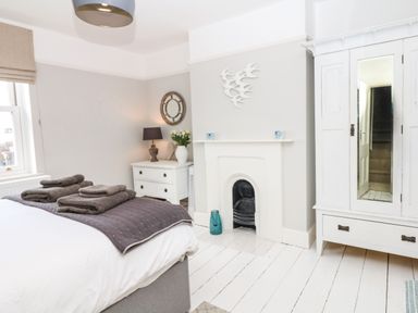 A bedroom with a bed holding folded towels a white dresser with a lamp and flowers a white fireplace and a white wardrobe with a mirror at Cool Stone Cottage Instow near Bideford
