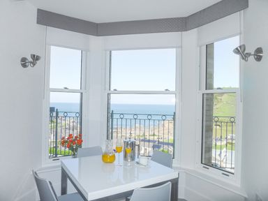 A dining area with a white table and chairs by three windows showing a sea view at 5 Arlington Villas in Ilfracombe