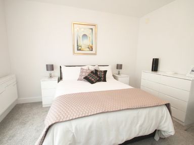 A bedroom with a bed a blanket pillows two bedside tables lamps a dresser and a framed picture on the wall at 5 Arlington Villas in Ilfracombe