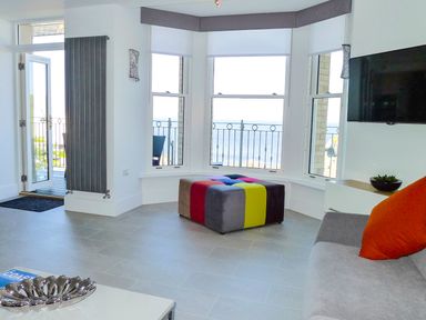 A living room with a sofa with an orange cushion a wall-mounted TV a colorful ottoman and windows overlooking the sea at 5 Arlington Villas in Ilfracombe