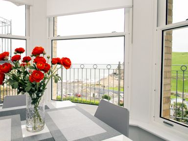 A table with a vase of red flowers in front of large windows overlooking a coastal view at 5 Arlington Villas in Ilfracombe