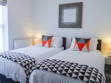 A bedroom with two single beds with patterned throws and pillows a bedside table with a lamp and wall art above the beds at 5 Arlington Villas in Ilfracombe