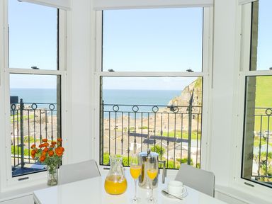 A dining area with a white table and chairs with flowers and orange juice in glasses by a window overlooking the sea at 5 Arlington Villas Ilfracombe
