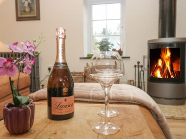 A wooden table with a bottle of Lanson champagne, two wine glasses, and a purple flower pot in a living room with a fireplace and window at Trevoole Barn in Praze An Beeble