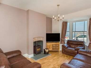 A living room with leather sofas a wood stove a television on a wooden stand and a window showing parked cars and water at The Crab Shell in Appledore near Bideford