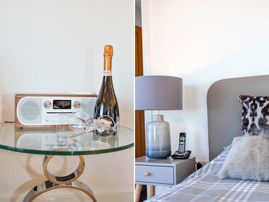A glass table with a radio, champagne bottle, and two glasses next to a bedside table with a lamp, cordless phone, and bed with pillows at Ocean Breeze in Ilfracombe