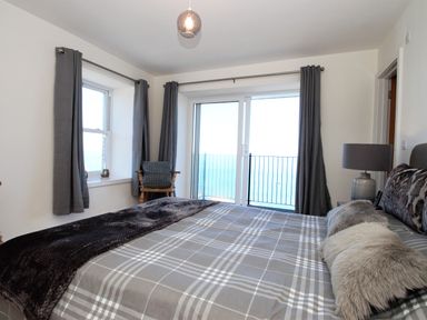 A bedroom with a bed covered in checkered bedding and fur pillows a lamp on a bedside table a wooden chair with a cushion and windows with gray curtains overlooking the sea at Ocean Breeze in Ilfracombe