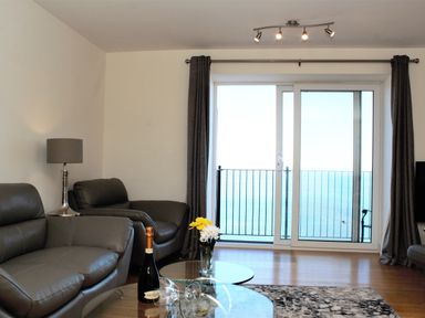 A living room with leather sofas a glass coffee table with a bottle and glasses and a balcony door showing the sea at Ocean Breeze in Ilfracombe