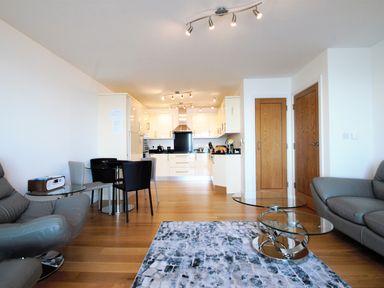 An open-plan living room and kitchen with grey leather chairs glass dining table and wooden floor at Ocean Breeze in Ilfracombe