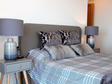 A bedroom with a bed with patterned pillows and a gray plaid bedspread between two bedside tables with lamps at Ocean Breeze in Ilfracombe