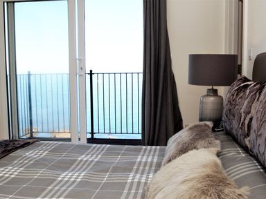 A bedroom with a bed covered with cushions and a lamp on a bedside table next to a balcony with a sea view at Ocean Breeze in Ilfracombe