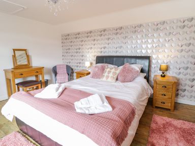 A bedroom with a double bed with pink and white bedding two wooden bedside tables with lamps a wooden dressing table a gray chair and heart patterned wallpaper at Tai Ucha in Cerrigydrudion