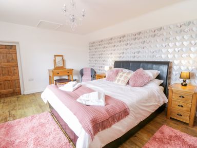 A bedroom with a bed with pink cushions and a pink throw blanket two wooden bedside tables with lamps a wooden door a wooden dressing table a chair and heart patterned wallpaper at Tai Ucha in Cerrigydrudion