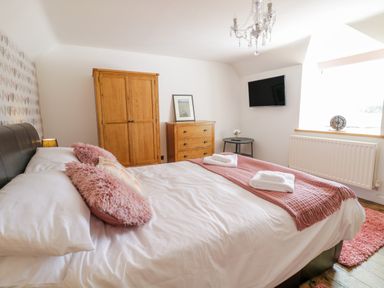 A bedroom with a bed with pink pillows and blanket a wooden wardrobe dresser a wall-mounted television and a window at Tai Ucha in Cerrigydrudion
