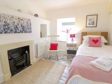 A bedroom with a single bed with pink blanket and heart pillow a white rocking chair with a red cushion a small nightstand with lamp flowers and framed picture at Tai Ucha in Cerrigydrudion