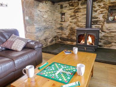A living room with a leather sofa a wooden coffee table with a scrabble game and mugs and a wood burning stove set in a stone wall at Tai Ucha in Cerrigydrudion
