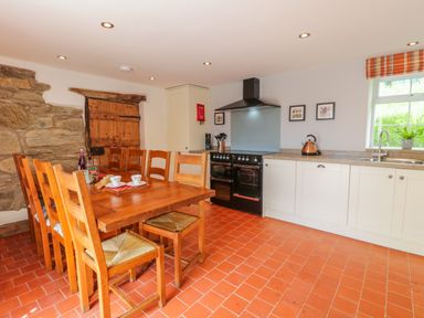 A kitchen with a wooden dining table and chairs a black stove white cabinets and a window at Tai Ucha in Cerrigydrudion