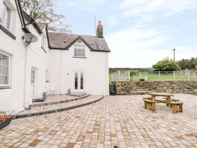 An outdoor patio area with paved flooring a wooden picnic table white painted house stone wall and steps leading to a fenced grassy yard at Tai Ucha in Cerrigydrudion
