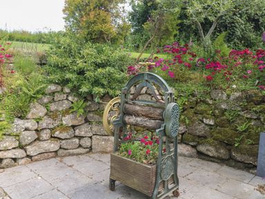 An outdoor area with a stone wall and plants including red flowers and greenery and an old fashioned garden roller with logs and a flower box at The Hayloft in St Neot near Dobwalls