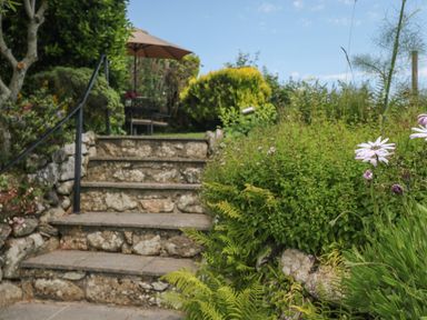 Stone steps leading up to a garden area with a table umbrella surrounded by green plants and flowers at The Hayloft in St Neot near Dobwalls