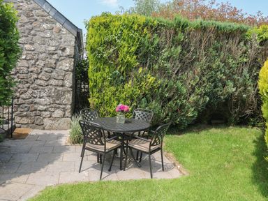 A patio with a round table and four chairs with flowers on the table next to a stone building and large bushes at The Hayloft in St Neot near Dobwalls