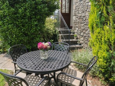 A metal outdoor table with four chairs and a vase of flowers near stone steps and green shrubs at The Hayloft in St Neot near Dobwalls