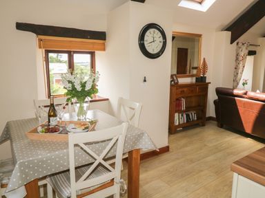 A dining area with a table set with food and flowers next to a window and a living room with a leather sofa and wooden furniture at The Hayloft in St Neot near Dobwalls