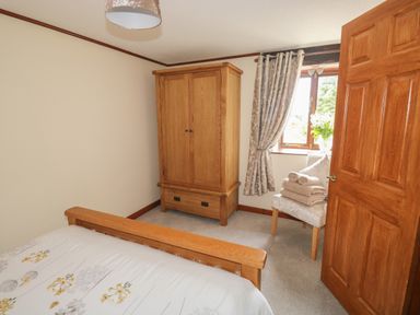 A bedroom with a wooden bed frame wooden wardrobe a chair with folded towels and a window with curtains at The Hayloft in St Neot near Dobwalls