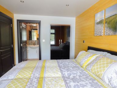 A bedroom with a bed covered by patterned yellow and gray bedding a black wardrobe two doors one open to a bathroom and a yellow wooden wall with a beach painting at Hidden Gem Cabin in Lettermacaward near Dungloe County Donegal
