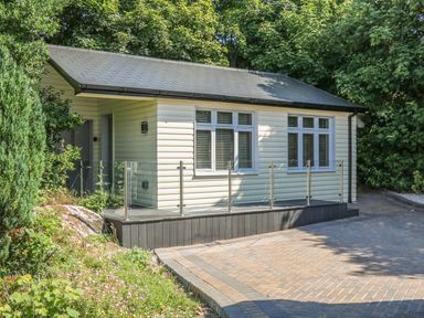 A small house with cream siding windows and a glass railing on a deck near a paved driveway and green trees at Hidden Gem Cabin in Lettermacaward near Dungloe County Donegal