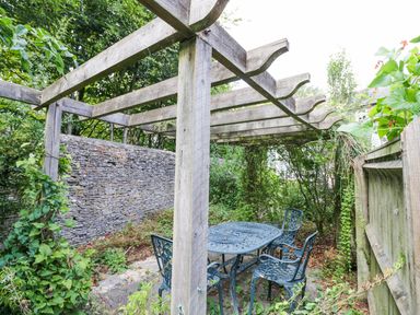 An outdoor seating area with a metal table and four chairs under a wooden pergola surrounded by plants and a stone wall at Blenheim Edge The Causeway in Woodstock